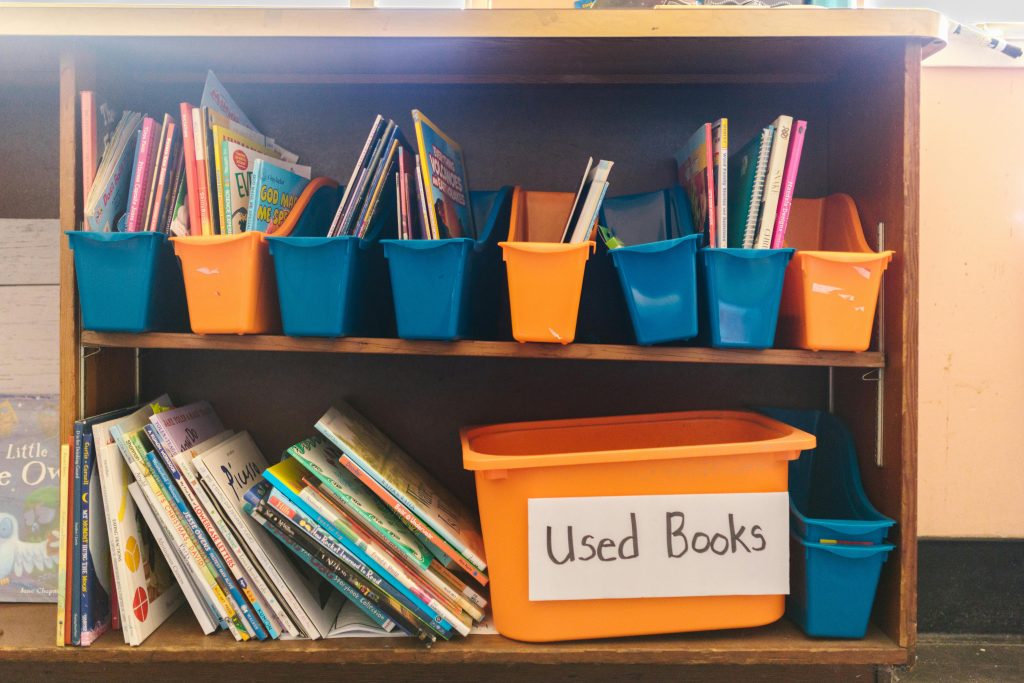 Colorful bins filled with children's books on a wooden shelf, labeled as 'Used Books'.